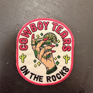 Op de afbeelding: Geborduurde patch met een witte achtergrond en een roze rand. De patch heeft de tekst "COWBOY TEARS ON THE ROCKS" in het rood. Een hand met rode nagels houdt een cocktail met een schijfje limoen vast. Groene en gele accenten.