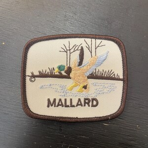 May include: Embroidered patch depicting a mallard duck in flight over water, with bare trees. The patch has a brown border and the word "MALLARD" in capital letters.