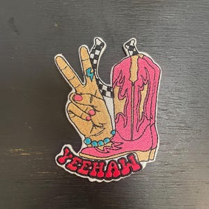 May include: Embroidered patch featuring a hand making a peace sign, a pink cowboy boot, and the word "Yeehaw" in red. The hand has a turquoise ring and a bracelet. The boot has a checkered pattern and a pink and yellow design.
