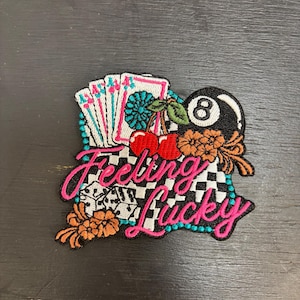 Può includere: Toppa ricamata a tema gioco d'azzardo. Presenta carte da gioco, dadi, una palla nera numero 8, ciliegie e fiori. La frase "Feeling Lucky" è scritta in corsivo rosa con un contorno turchese, su sfondo a scacchi bianco e nero.