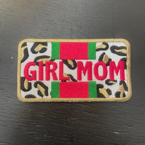 May include: Embroidered patch with the text "GIRL MOM" in red block letters. The patch has a white background with a leopard print pattern. Red and green stripes are above and below the text, and the border is gold.