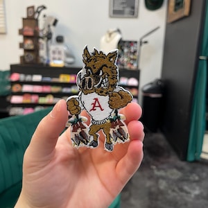 Puede incluir: Parche bordado con una mascota de jabalí de dibujos animados en marrón, blanco y rojo. El parche representa al jabalí con una expresión feroz, vistiendo una camisa blanca con una "A" roja y sosteniendo dos paquetes.