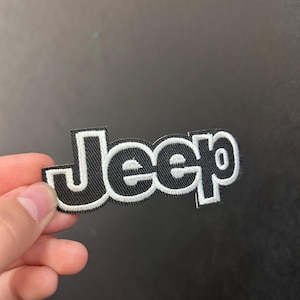 May include: Black and white embroidered patch featuring the word "Jeep" in a bold, stylized font. The patch has a black background with white stitching outlining the letters. The patch is held by a hand.
