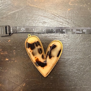 May include: A heart-shaped pendant featuring a cream and brown tortoiseshell pattern encased in a gold-toned metal frame. A black measuring tape is positioned above the pendant, showing it measures approximately 2.25 inches in width.
