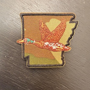Peut inclure: Écusson brodé représentant un canard en vol avec des plumes orange et blanches, une tête verte et un bec brun. L'écusson a la forme de l'État de l'Arkansas, avec un fond camouflage et une bordure noire.