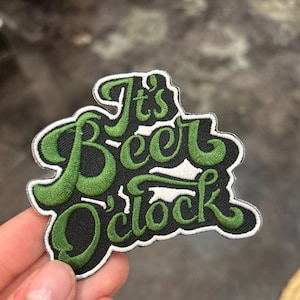 Puede incluir: Parche bordado con la frase "It's Beer O'clock" en verde sobre fondo negro. El parche tiene un borde blanco y está diseñado para ser cosido o planchado en tela.