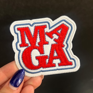 May include: Embroidered patch featuring the letters "MGA" in red, with blue and white detailing. The patch has a white background and a blue border. The patch is held by a person with dark blue nail polish.