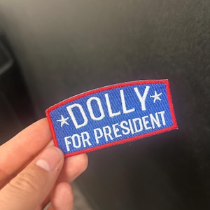 Peut inclure: Un écusson rectangulaire bleu avec le texte "DOLLY FOR PRESIDENT" en lettres blanches. L'écusson a une bordure rouge et deux étoiles blanches. L'écusson est tenu par une main.
