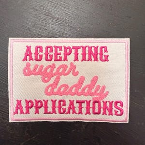May include: Embroidered patch with the text "ACCEPTING sugar daddy APPLICATIONS" in pink lettering on a white background. The patch has a pink border and is rectangular in shape. The patch is likely for clothing or accessories.