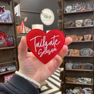 Peut inclure: Un écusson rouge en forme de cœur avec l'inscription "Tailgate Season" brodée en blanc. L'écusson est présenté devant un présentoir de magasin avec divers autocollants et marchandises.