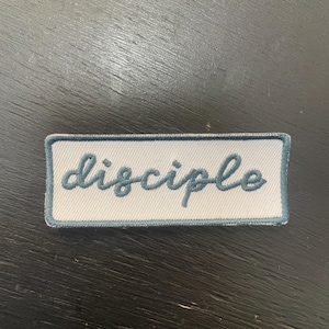 May include: A rectangular patch with the word "disciple" embroidered in cursive teal lettering. The patch has a white background with a teal border. The patch is likely for clothing or accessories.