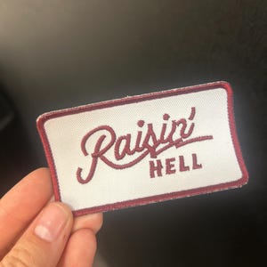 Peut inclure: Écusson brodé avec les mots "Raisin' HELL" en fil bordeaux sur fond blanc. L'écusson a une bordure bordeaux et est de forme rectangulaire. L'écusson est tenu par une personne.