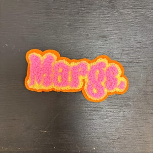 Peut inclure: Un écusson en chenille rose et jaune avec le nom "Marge" dans une police rétro. L'écusson a une bordure orange et est posé sur une surface en bois sombre.