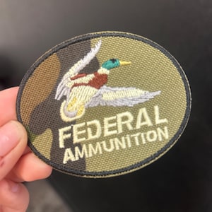 Peut inclure: Écusson brodé de forme ovale avec un motif camouflage. Il représente un canard en vol avec une tête verte, une poitrine marron et des ailes blanches. Le texte "FEDERAL AMMUNITION" est brodé en blanc.