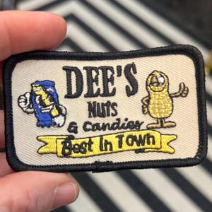 May include: A white and black embroidered patch with the text "DEE'S NUTS & CANDIES Best In Town". The patch features a cartoon peanut and a cartoon can of soda.