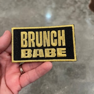 May include: Black rectangular patch with gold embroidered text that reads "BRUNCH BABE". The patch has a gold border and is held in a hand. The patch is likely an accessory for clothing or bags.