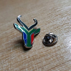 Puede incluir: Un pin de plata con un diseño de cabeza de antílope en verde, amarillo, rojo y azul. El pin está sobre una superficie de madera.