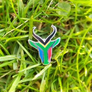 May include: A metal pin shaped like an antelope head with the colors of the South African flag: green, yellow, black, red, and blue.