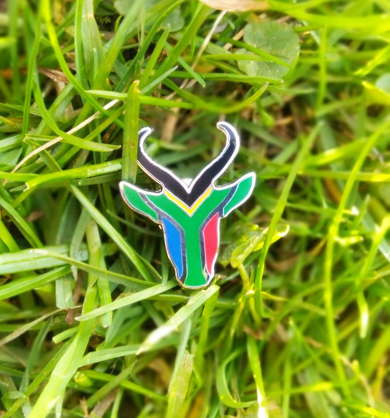 May include: A metal pin shaped like an antelope head with the colors of the South African flag: green, yellow, black, red, and blue.