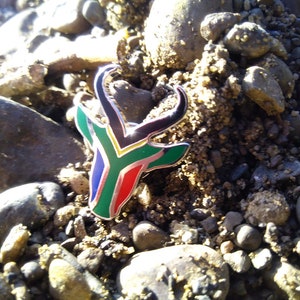 May include: A silver pin with a black, green, yellow, and blue antelope design. The antelope is a symbol of South Africa and the colors represent the South African flag.