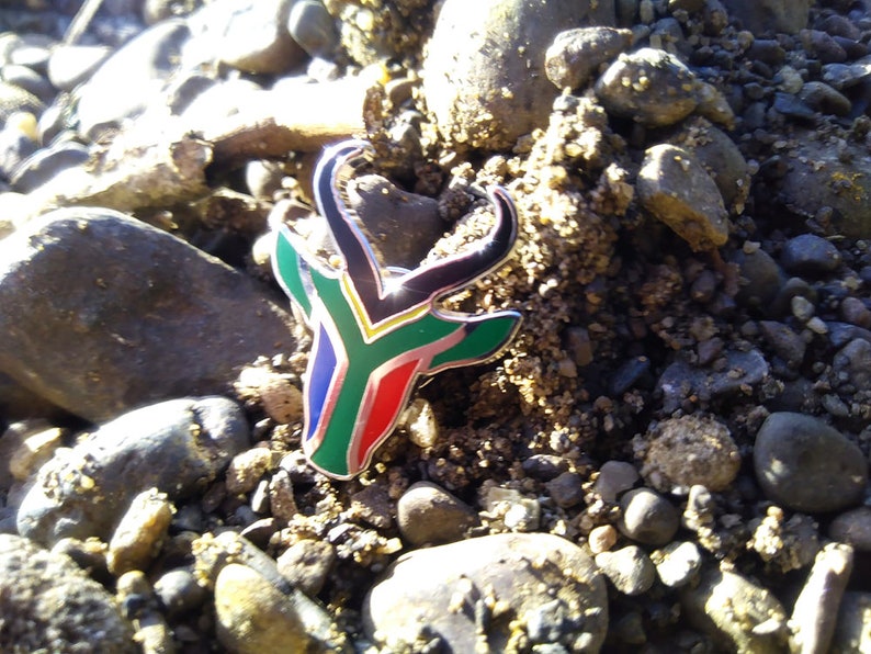 May include: A silver pin with a black, green, yellow, and blue antelope design. The antelope is a symbol of South Africa and the colors represent the South African flag.