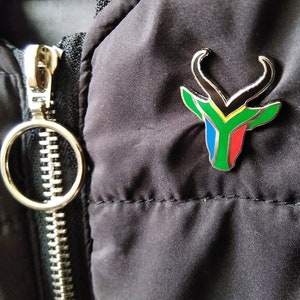 May include: A silver pin with a black, green, yellow, blue, and red springbok antelope design. The pin is attached to a black jacket.