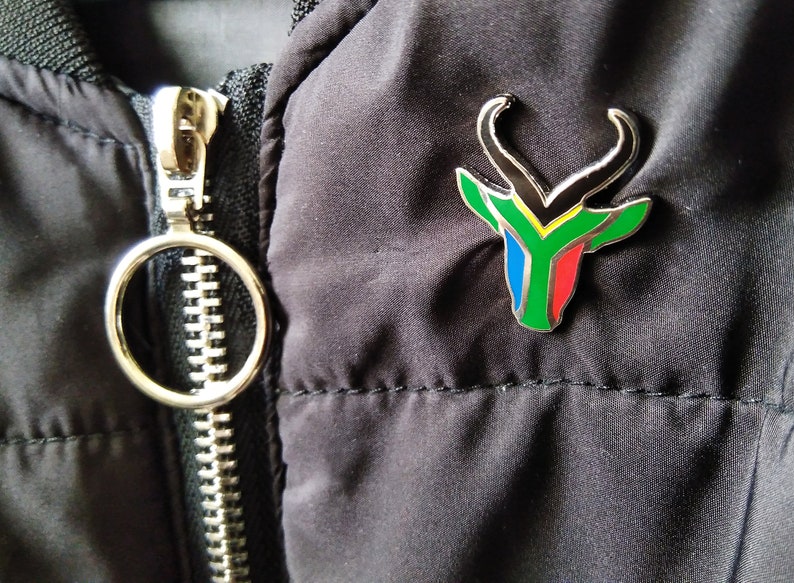 May include: A silver pin with a black, green, yellow, blue, and red springbok antelope design. The pin is attached to a black jacket.