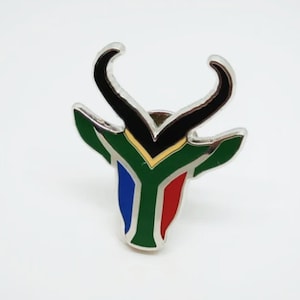 May include: A silver metal pin with a black springbok antelope head design. The antelope's head is colored in the colors of the South African flag: green, gold, blue, and red.