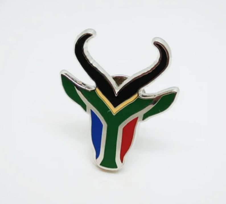 May include: A silver metal pin with a black springbok antelope head design. The antelope's head is colored in the colors of the South African flag: green, gold, blue, and red.