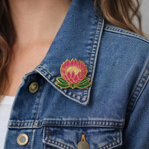 May include: A gold-toned metal enamel pin shaped like a protea flower with vibrant pink petals, a textured cream-coloured centre, and dark green leaves at the base, attached to the collar of a blue denim jacket with visible metal buttons.