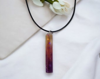 Puede incluir: Un collar de cordón negro con un colgante rectangular largo. El colgante está hecho de resina transparente con un degradado de colores amarillo, naranja y morado. Hay pequeñas partículas brillantes incrustadas en la resina.