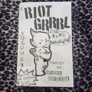 Puede incluir: Un fanzine en blanco y negro titulado "A Riot Grrrl Zine" con el texto "Punk Bubblegum" en un bocadillo. La portada presenta un personaje de dibujos animados y el texto "Volume 3" y "Featuring: The Venomous Pinks". Creado por Paige Daniel.