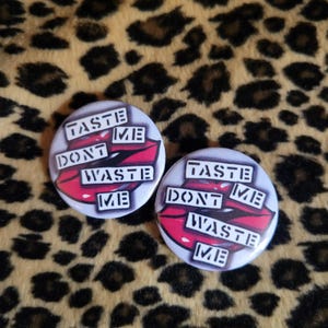 May include: Two round white buttons with red lips and black text that reads "Taste Me Don't Waste Me".