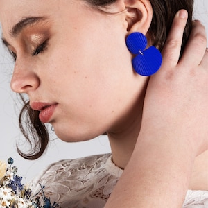 May include: A woman wearing a white lace top with blue geometric earrings. The earrings are made of two overlapping circles with a textured surface.