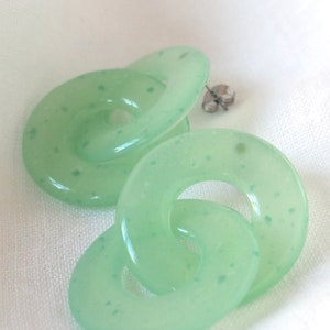 May include: A pair of light green, interlocking circle earrings. The earrings have a translucent appearance with small, darker green speckles throughout. The earring posts are silver-colored.