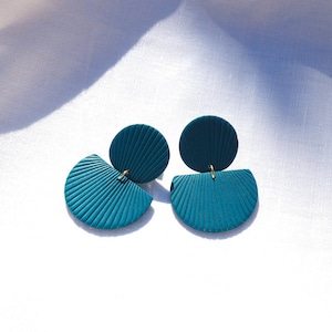 May include: A pair of teal-colored earrings with a textured, fan-shaped design. Each earring features a circular top and a half-circle bottom, connected by a small gold-colored loop. The earrings are displayed on a white, textured surface.