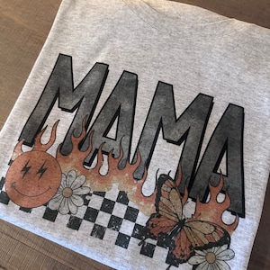 以下が含まれることがあります： 炎と蝶の模様が施された、黒い文字で「MAMA」とプリントされたグレーのTシャツです。