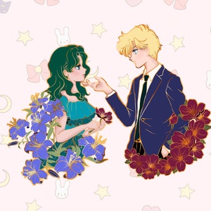Op de afbeelding: Een gouden emaille pin met een stel in een romantische pose. De vrouw heeft groen haar en draagt een blauwe jurk. De man heeft blond haar en draagt een blauw pak. Beide zijn omringd door paarse en rode bloemen.
