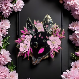 May include: A black and gold deer head pin, adorned with pink floral accents and surrounded by a wreath of pink flowers. The pin features a crescent moon and star details, creating a whimsical and elegant design.