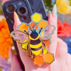 Puede incluir: Un soporte para anillo de teléfono de abeja dorada con un diseño de panal. La abeja tiene rayas negras y amarillas y alas transparentes. El panal es amarillo y naranja con una textura brillante.