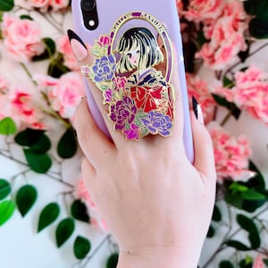 Könnte beinhalten: Eine goldfarbene Handyhülle mit einem violetten Blumendesign. Das Design zeigt eine Frau mit schwarzen Haaren und einem rot-goldenen Kimono. Der Text "Shadow Stamina" ist in einem Kreis um die Frau geschrieben.