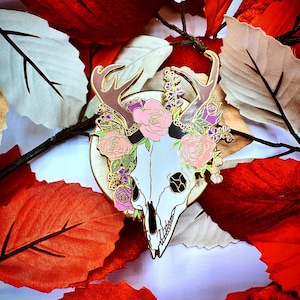 May include: A gold-tone enamel pin featuring a white deer skull with antlers, surrounded by pink, purple, and green flowers. The pin is set against a background of red and white leaves.