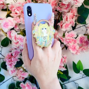 Può includere: Una custodia per telefono color lavanda con un portachiavi Sailor Moon dorato. Il portachiavi presenta un'illustrazione dettagliata di Sailor Moon nella sua posa iconica con uno sfondo scintillante.