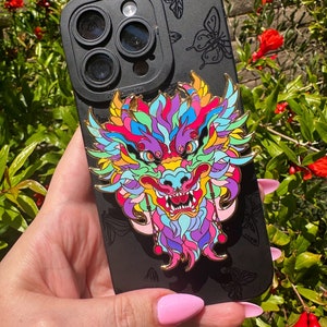 Puede incluir: Una funda de teléfono negra con un diseño de dragón colorido. El dragón está hecho de esmalte y tiene detalles dorados. La funda del teléfono tiene una cubierta de lente de cámara con tres lentes.