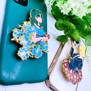 Op de afbeelding: Twee telefoonhoesjes met decoratieve emaille-pins met anime-personages. De eerste hoes is groen met een blauwe en gouden pin van een vrouw met lang groen haar. De tweede hoes is transparant met een rode en gouden pin van een man met blond haar.