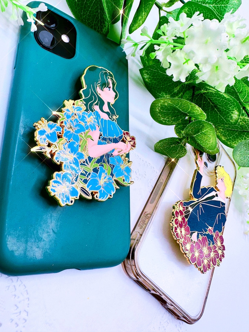 Op de afbeelding: Twee telefoonhoesjes met decoratieve emaille-pins met anime-personages. De eerste hoes is groen met een blauwe en gouden pin van een vrouw met lang groen haar. De tweede hoes is transparant met een rode en gouden pin van een man met blond haar.