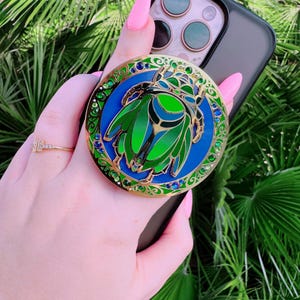May include: A gold-toned phone grip with a blue and green jeweled scarab beetle design. The grip is attached to a black phone case.