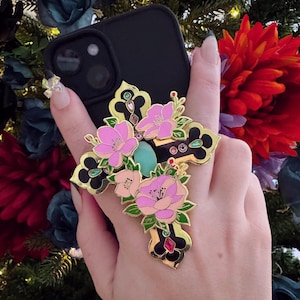 May include: Black phone case with a decorative cross-shaped phone grip. The grip features pink floral designs, gold accents, and a turquoise center stone. The phone case is held in front of a backdrop of colorful flowers.