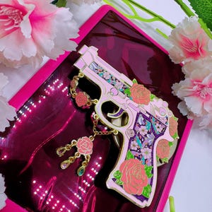 May include: A pink decorative pistol pin with floral accents, including roses and a chain with dangling charms. The pin features a purple and floral design on the handle and is set against a pink background.