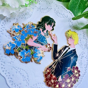 Op de afbeelding: Twee gouden emaille-pins die een stel voorstellen. De vrouw draagt een blauwe jurk en heeft groen haar. Ze is omringd door blauwe bloemen. De man draagt een zwart pak en is omringd door rode bloemen.